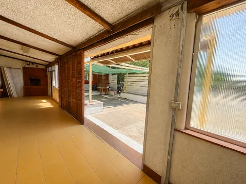 Ambiente coperto con pavimento in piastrelle gialle e soffitto a travi di legno, chiuso su un lato da persiane in legno; una grande apertura dà sulla corte esterna, riparata da un telo verde.