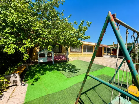 Giardino con prato sintetico all'ombra di un grande albero: un'altalena in metallo verde in primo piano, una casetta gioco e una panca addossate al muro, e il fronte dell'edificio con le vetrate sul fondo.