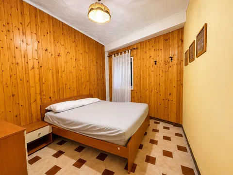 Camera da letto con due pareti rivestite in perlinato di legno chiaro: letto matrimoniale con testiera in legno, comodino e comò, pavimento a scacchi bianco e marrone, finestra con tenda bianca.