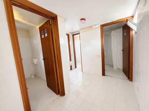 Blocco dei servizi igienici: disimpegno piastrellato in bianco con più porte in legno, due contrassegnate dai pittogrammi dei bagni, una doccia con tenda sulla destra e un rilevatore a soffitto.