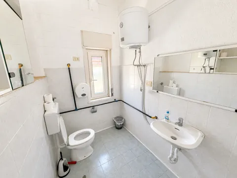 Bagno interamente piastrellato di bianco, con scaldacqua elettrico a parete, vaso, lavabo sospeso, specchio a tutta larghezza e una finestrella alta.