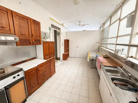 Cucina di servizio in linea: pensili e basi in legno scuro, cappa in acciaio, doppio lavello sotto una parete vetrata a vetri retinati, un tavolo da lavoro al centro e la porta di uscita di sicurezza segnalata sul fondo.