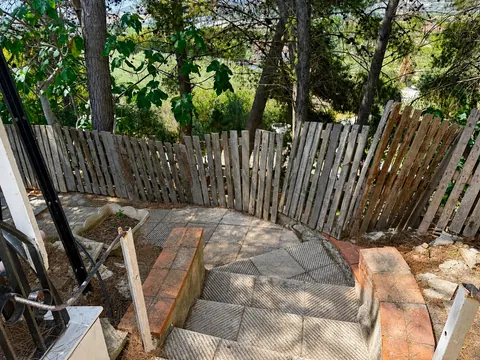 Scala esterna in discesa verso una piccola corte pavimentata a mosaico di cemento, chiusa da una staccionata di doghe grezze; oltre la staccionata, pini e alberi di fico.