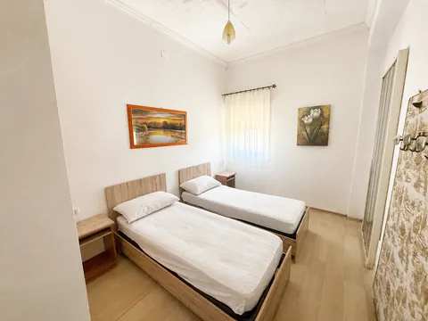 Camera a due letti singoli con testiere in legno chiaro e comodini abbinati, pavimento in laminato, due quadri alle pareti bianche e una finestra con tenda leggera.