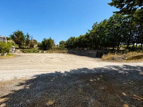 Ampio piazzale sterrato compattato, delimitato sul fondo da un muretto in pietra e da un filare di pini; sulla sinistra un piccolo chiosco e la vegetazione del bordo.