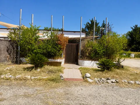 Ingresso pedonale al retro del fabbricato: cancello in legno fra due fioriere circolari, sei pali metallici verticali di una struttura, arbusti cresciuti liberamente e un vialetto piastrellato che parte dalla ghiaia.