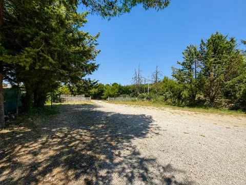 Spiazzo in ghiaia all'ombra di grandi conifere, chiuso sul fondo da una staccionata in legno; sulla destra il terreno prosegue aperto verso la campagna.