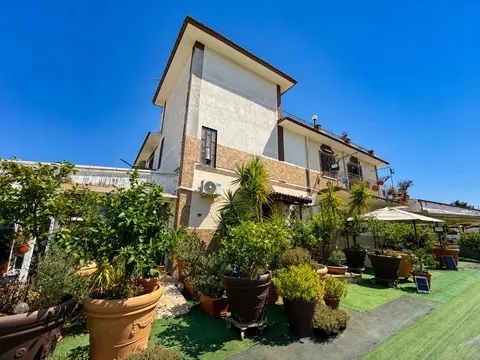 La villa vista dal giardino sotto un cielo terso: volume su due livelli con fascia in pietra e parapetto della terrazza in alto, agrumi e piante grasse in grandi vasi di terracotta, prato sintetico e ombrelloni bianchi sulla destra.
