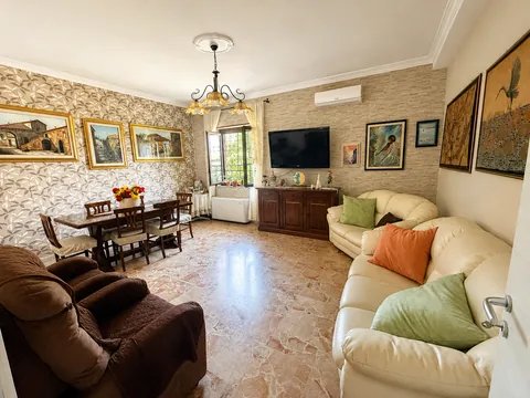 Soggiorno con pavimento in marmo rosato: due divani in pelle chiara con cuscini colorati, tavolo da pranzo in legno con sei sedie, credenza, televisore a parete e quadri su carta da parati a foglie.