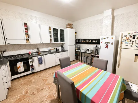 Cucina abitabile con mobili bianchi lungo due pareti, piano cottura a gas, forno, due frigoriferi affiancati e tavolo con tovaglia a righe colorate e quattro sedie.