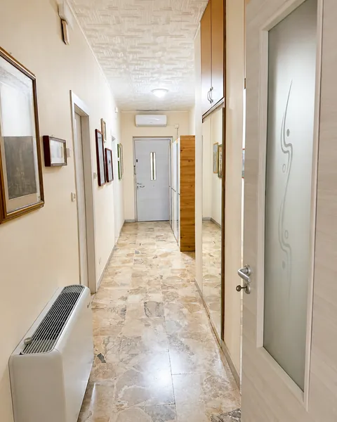 Corridoio interno con pavimento in marmo chiaro venato, quadri incorniciati lungo la parete di sinistra, un ventilconvettore a pavimento e, in fondo, la porta blindata d'ingresso.