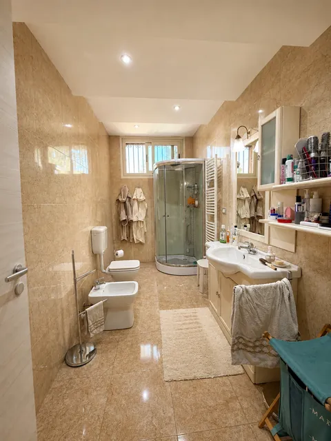 Bagno rivestito in marmo beige dal pavimento al soffitto, con box doccia angolare in cristallo, vaso e bidet, mobile lavabo con specchio e scaldasalviette a parete.