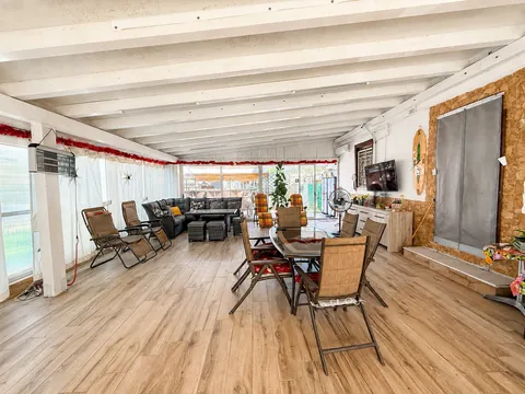 Veranda coperta con travi bianche a vista e pavimento in gres effetto legno: divano angolare scuro, tavolo da pranzo con sedie pieghevoli, tende chiare lungo le vetrate e parete in pietra sulla destra.