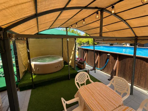 Area esterna sotto un gazebo a volta con telo e catena di lampadine: vasca idromassaggio circolare su prato sintetico, piscina fuori terra con rivestimento in doghe di legno e tavolo con sedie bianche in primo piano.