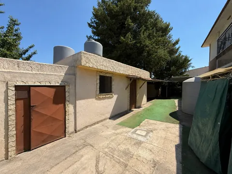 Corte di servizio pavimentata in cemento: corpo basso con portone in ferro, due serbatoi idrici cilindrici sulla copertura e un terzo a terra, cipresso alto sul fondo.