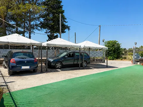 Area di sosta con due automobili all'ombra di tre grandi ombrelloni bianchi, muro di recinzione in blocchi di calcestruzzo, pini sullo sfondo e prato sintetico verde in primo piano.