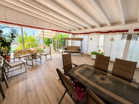 Interno della veranda con travi bianche: dondolo a tre posti, divanetto a fiori, poltroncine e tavolo da pranzo con piano in vetro, tende bianche lungo tutto il perimetro e stufa radiante appesa.