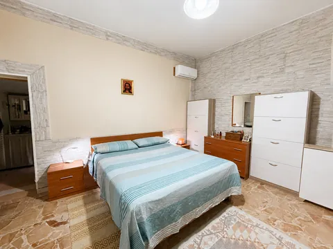 Camera matrimoniale con due pareti rivestite in pietra ricostruita chiara: letto con testiera in legno, comodini, comò con specchio e cassettiera bianca, pavimento in marmo.